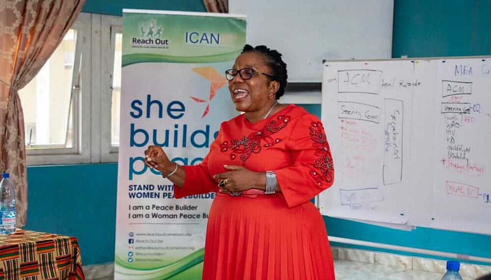 A picutre of Esther Omam, Executive Director of the organisation "Reach out" from Cameroon, speaking at a workshop. Esther wears a red dress and black glasses.